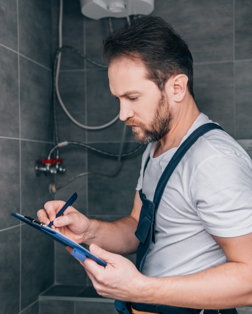 adult male plumber writing in clipboard and checking electric boiler in bathroom